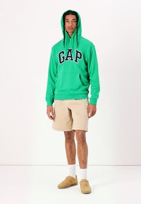 Felpa verde con logo "GAP", in materiale misto cotone, tasca frontale, abbinata a pantaloni corti beige e sneakers color sabbia. Modello in piedi su uno sfondo bianco.