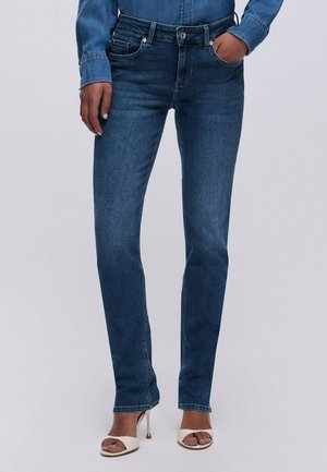 Osoba nosząca dopasowane niebieskie dżinsy z przednimi i tylnymi kieszeniami, w połączeniu z białymi sandałami na wysokim obcasie i pasującą niebieską koszulą dżinsową.