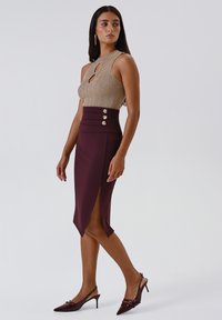 Donna che indossa un top senza maniche beige in maglia e una gonna a tubino bordeaux a vita alta con spacco frontale e dettagli in bottoni, abbinata a scarpe col tacco bordeaux.
