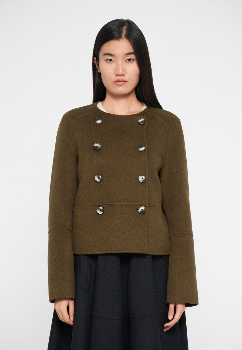 Oliivroheline kahekordse nööpimisega villane jakk, millel on suured mustad nööbid ja laiad varrukad, koos volditud musta seelikuga.