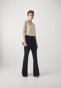 Top lungo a maniche corte con paillettes dorate, pantaloni neri svasati, in mano una pochette nera, texture liscia, design aderente, linee pulite.