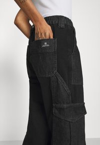 Black denim cargo pants with a soft texture, featuring multiple pockets and visible stitching details. Elastic waistband for adjustable fit.