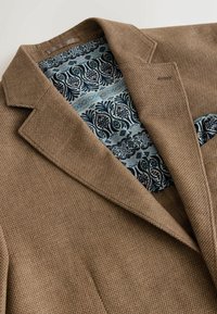 Blazer marron texturé avec une doublure intérieure fleurie bleue et noire visible le long du col et du bord de la poche.