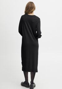 Lång svart tröjaklänning med långa ärmar, rund halsringning och rak passform. Texturen verkar slät; bärs med svart fotbeklädnad.