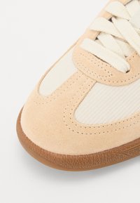 Baskets en daim beige avec semelle en caoutchouc texturée, ornées de détails en tissu clair et de lacets plats. Bout arrondi avec coutures détaillées.