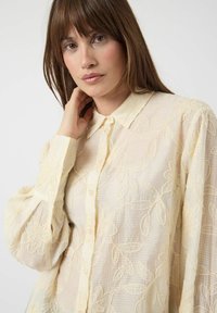 Krämfärgad blus med blomsterbroderi, texturerad väv, knappstängning, spetskrage och samlade ärmar. Detaljerade löv- och blommönster.