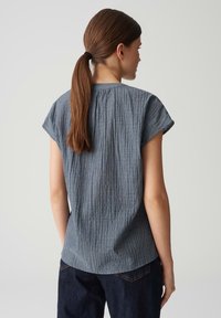 Blusa grigia con texture, maniche corte, caratterizzata da una piega sul retro e scollatura rotonda, abbinata a jeans in denim scuro.