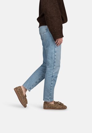 Personne portant un jean bleu clair, un pull marron et des mocassins imprimés léopard avec des glands, debout devant un fond uni.