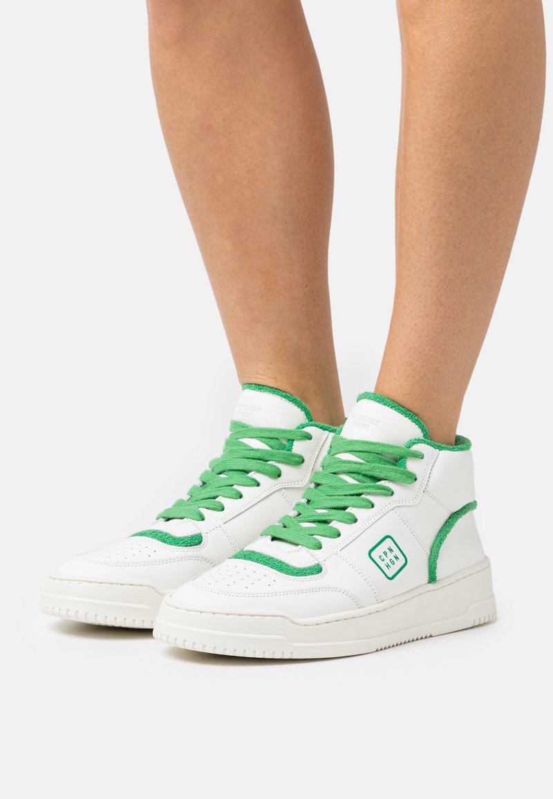 Baskets montantes blanches avec des lacets et des bordures verts portées sur des jambes nues, avec un écusson carré du logo sur le côté extérieur.