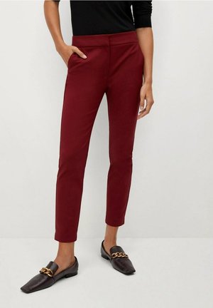 Persoon draagt nauwsluitende bordeauxrode broek en zwarte loafers met gouden kettinkje, hand in linkerzak, staand tegen een effen achtergrond.