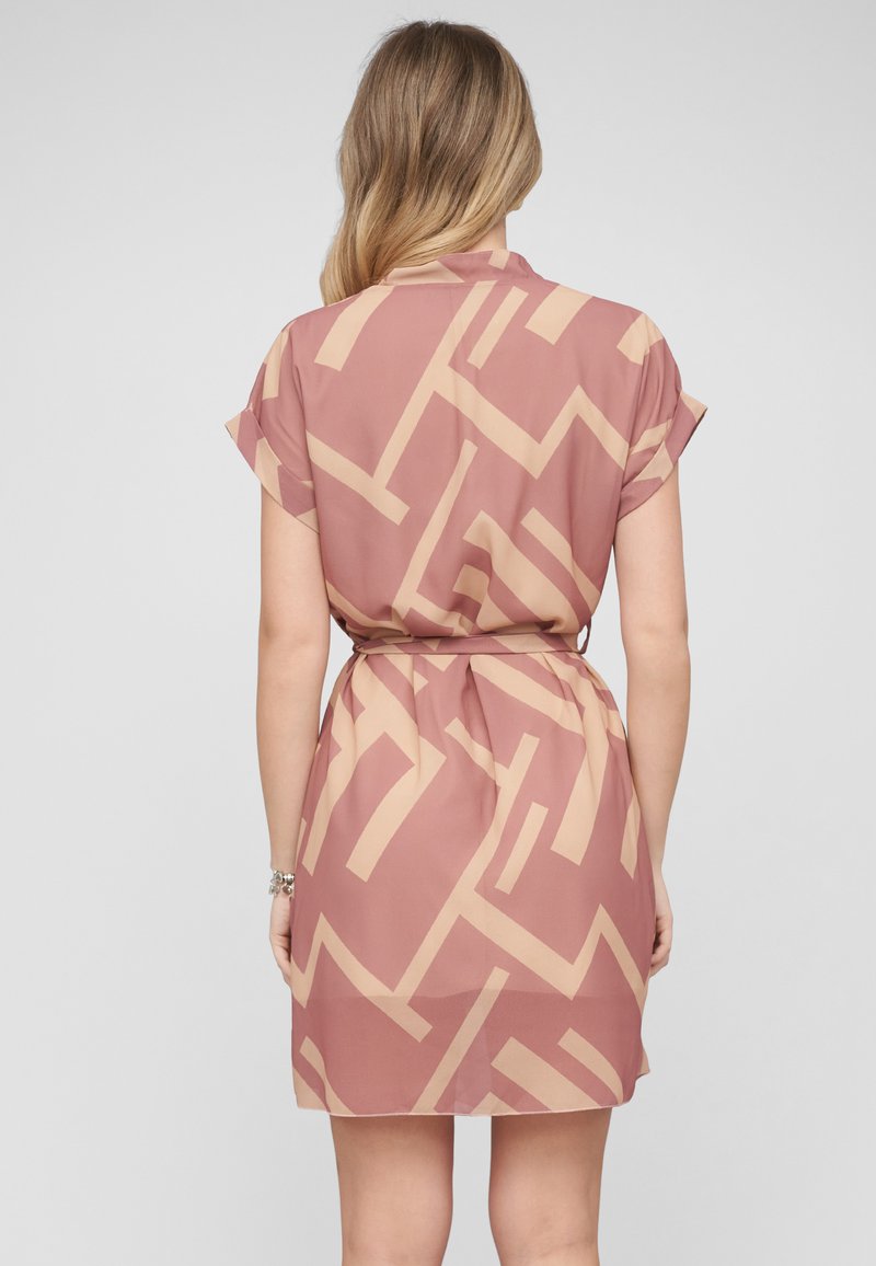 Femme aux longs cheveux blonds portant une robe courte à manches courtes, longueur genoux, avec un motif géométrique rose et beige, vue de dos.