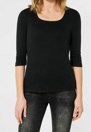 Femme portant un haut noir ajusté à encolure dégagée avec manches trois-quarts et un jean noir délavé, debout les bras détendus le long du corps.