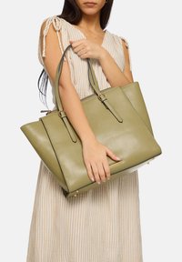 Sac fourre-tout en cuir vert avec une forme structurée, deux poignées robustes et des accents en métal doré. Texture lisse avec des coutures contrastées.