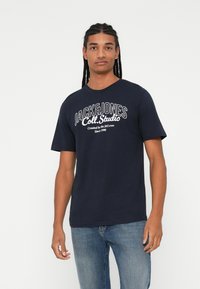 Tumesinine puuvillane T-särk lühikeste varrukatega, ümmargune kaelus ja ees valge trükitud tekst "JACK&JONES Coll.Studio".