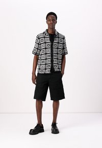 Chemise à manches courtes à motifs noir et blanc avec un design géométrique, associée à un short cargo noir et des chaussures lacées noires.