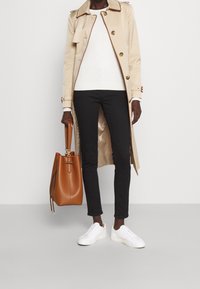 Person som bär en beige trenchcoat, vit tröja, svarta byxor och vita sneakers, som håller en stor brun läderhandväska.