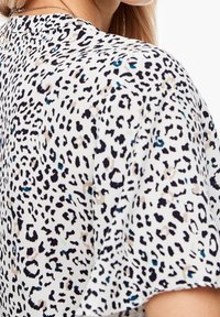 Kortärmad blus i ett lätt material med en vit bas och svart och beige leopardmönster, accentuerad med tealprickar.