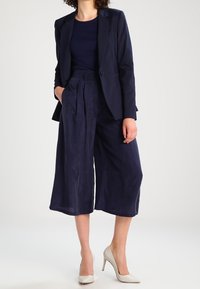 Námořnický blazer a odpovídající culottes z hladké tkaniny; přiléhavý střih se širokými nohavicemi; spárováno se světlými vysokými podpatky.