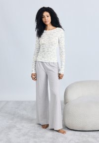 Haut léger à manches longues avec un motif floral bleu sur fond blanc cassé. Pantalon gris à jambes larges en matière douce. Modèle debout sur un tapis moelleux.