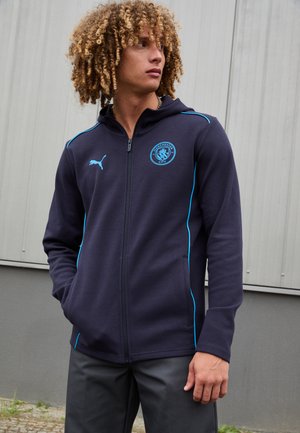 Sudadera con cremallera en color azul marino con acentos turquesa, que presenta un logo en el pecho, bolsillos tipo canguro y un diseño ajustado. Textura de tela suave.