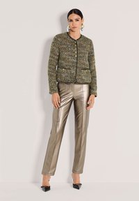 Frau in einer strukturierten grünen Knopfjacke aus Tweed, metallisch bronze-farbenen geraden Hose, schwarzen spitzen Schuhen und geometrischen Ohrringen stehend.