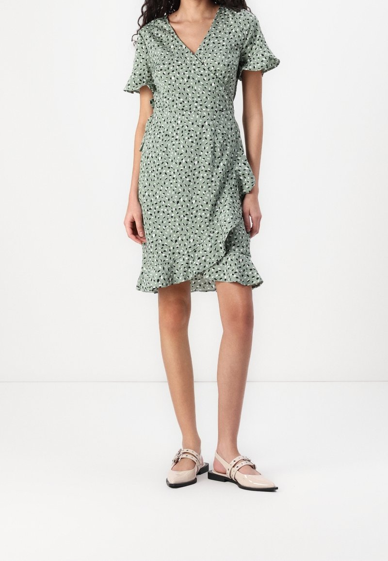 Femme portant une robe portefeuille à manches courtes vert clair avec des taches abstraites noires et blanches et un ourlet à volants, associée à des chaussures plates beiges.