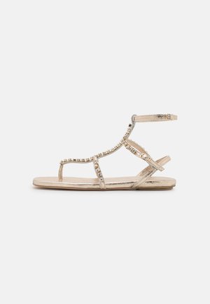 Sandalias planas metálicas doradas con múltiples correas, adornadas con brillantes de strass transparentes a lo largo del centro. Correa ajustable en el tobillo para un mejor ajuste.