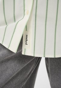 Chemise blanche à boutons avec des rayures verticales vertes et bleu marine, portant une étiquette "imbibée" sur l'ourlet. Assortie à un pantalon gris texturé.
