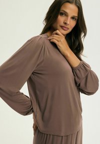 Długi rękaw w miękkim brązowym materiale, z bufiastymi rękawami, luźnym krojem i okrągłym dekoltem. Akcenty obejmują marszczone mankiety.