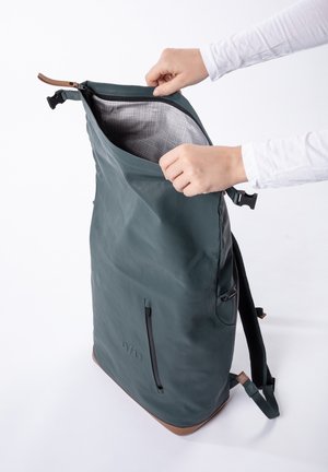 Handen openen een grote donkergroene rugzak met een zwarte ritssluiting en een bruine onderkant, waarbij het lichtgrijs geruite binnenvoering zichtbaar is.