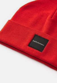 Red knit beanie with a folded cuff, featuring a square black patch labeled "Peak Performance." Soft texture and seamless design.