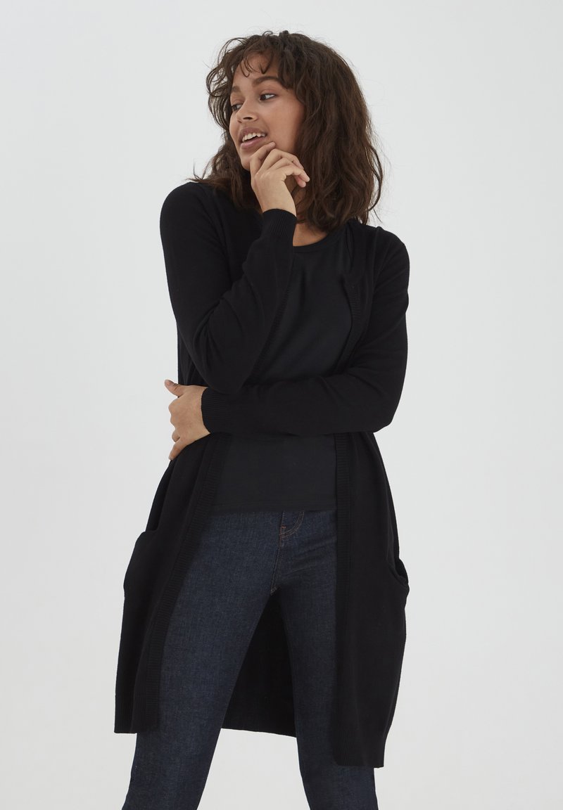 Zwarte lange cardigan met open voorkant, geribde manchetten en grote zijzakken, gedragen over een zwarte top en gecombineerd met donkere jeans. Gladde textuur.