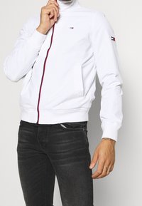 Chaqueta bomber blanca con cuello alto, con cremallera frontal con detalle rojo y pequeño logo en el pecho y la manga. Vaqueros negros puestos.