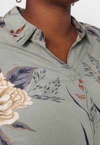 Kukkamekko vaaleanvihreästä kankaasta, jossa on beigejä ja purppuroita kuvioita. Varustettu kauluskauluksella ja nappiyksityiskohdilla. Pehmeä pinta.