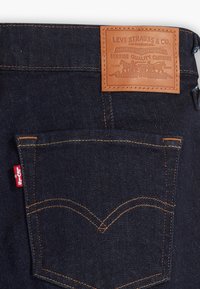 Tummansiniset farkut oransseilla ompeleilla, joissa on nahkaleima, johon on kaiverrettu "Levi Strauss & Co." ja punainen lappu takataskussa.