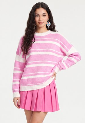 Jonge vrouw met lang donker haar, gekleed in een roze-wit gestreepte trui, een roze geplooide rok en kleurrijke lolly-oorbellen tegen een witte achtergrond.