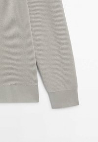 Manche d'un pull en tricot gris clair avec bord côtelé et motif de points verticaux texturés sur fond blanc.