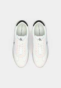 Stel sneakers van Calvin Klein in wit en beige met zwarte accenten, veters gestrikt, van bovenaf getoond op een witte achtergrond.