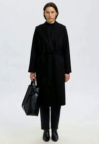 Mulher com um casaco preto cinto, calças pretas e botas pretas, segurando uma grande bolsa preta, de pé contra um fundo liso claro.