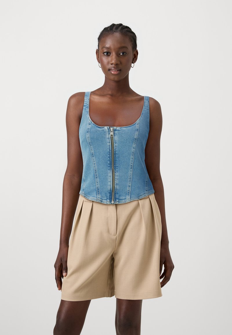 Top corsetto in denim con zip frontale, dotato di spalline larghe e silhouette aderente. Abbinato a pantaloni corti plissettati di un leggero beige.