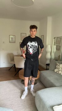 Černé grafické tričko s bílým a červeným designem; černé kraťasy; bílé tenisky; pozadí se stolem a židlemi; světlý koberec.