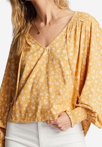 Blouse florale jaune avec un col en V, des manches froncées et des poignets élastiques, en tissu léger avec une texture douce et un léger éclat.