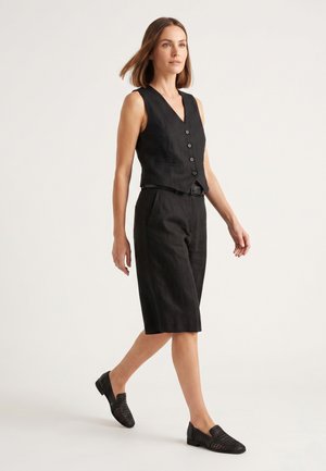 Kvinde iført sort ærmeløs knapvest, knælange sorte shorts og sorte vævede slip-on sko mod en ensfarvet lys baggrund.