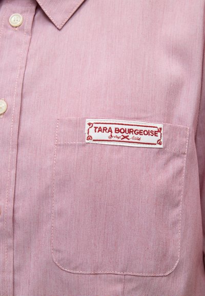Gros plan d'une chemise rose clair à col avec des boutons blancs et une poche portant une étiquette blanche et rouge "Tara Bourgeoise".