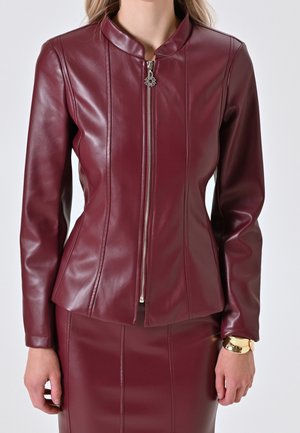 Femme portant une veste en cuir bordeaux ajustée avec une fermeture éclair à l'avant et une jupe assortie, debout, les bras détendus le long du corps.