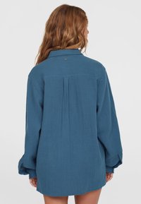 Blauwe button-up shirt gemaakt van gestructureerde stof met lange mouwen, een kraag en een rugplooi voor extra bewegingsvrijheid. Subtiel merkdetail op de nek.