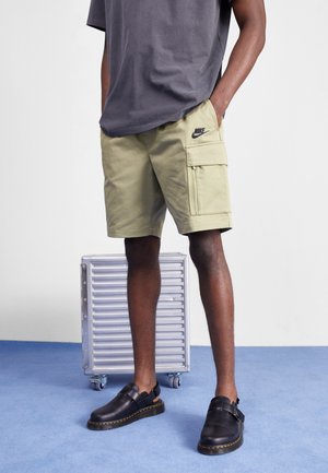 Man wearing beige Nike cargo shorts and black slip-on shoes standing next to a silver rolling suitcase on blue floor.