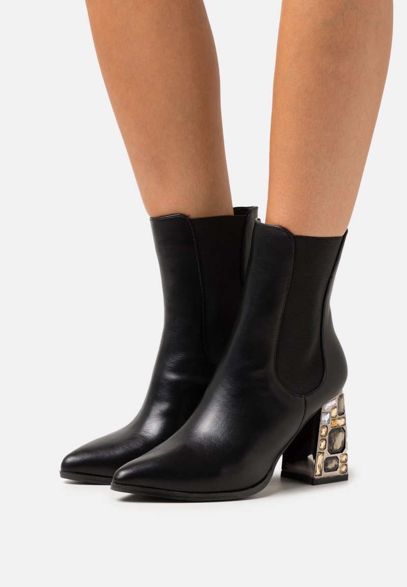Bottines en cuir noir avec bouts pointus et talons carrés ornés d'embellissements géométriques dorés et argentés, portées sur des jambes nues.
