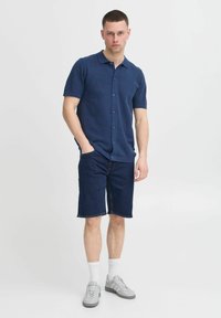 Polo shirt navy à manches courtes avec un bouton à l'avant, associé à un short en denim. Le modèle porte des baskets grises et des chaussettes blanches. Design simple et décontracté.