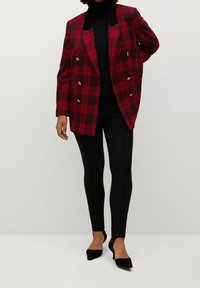 Frau in einem rot-schwarz karierten, doppelt-knöpfbaren Blazer über einem schwarzen Rollkragenpullover, schwarzen Leggings und schwarzen spitzen High Heels.